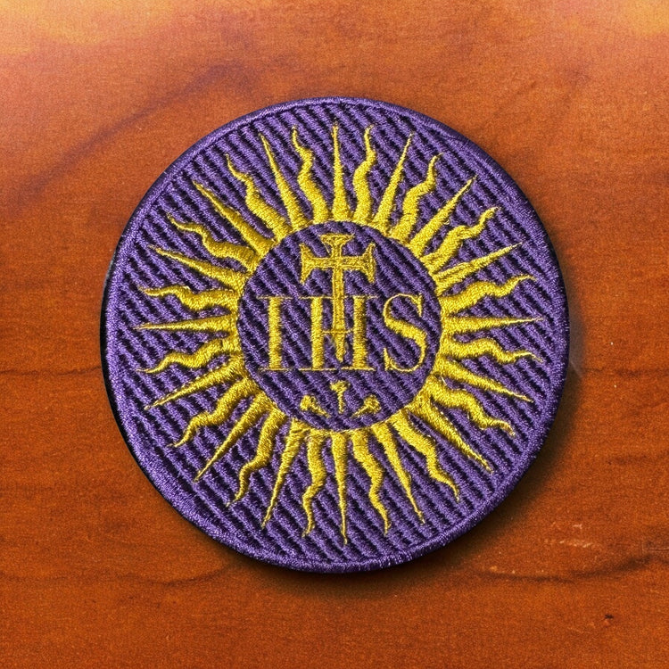 A purple and yellow round embroidered patch with IHS, or the Holy Name of Jesus monogram.